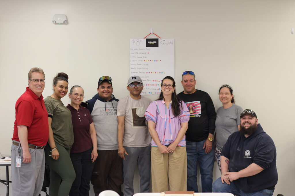 A diverse group of adult learners from Bradford White pose with their instructor from the Michigan Language Center after completing a weekly workplace ESL class. The chart on the wall shows a star-based student satisfaction reward system.